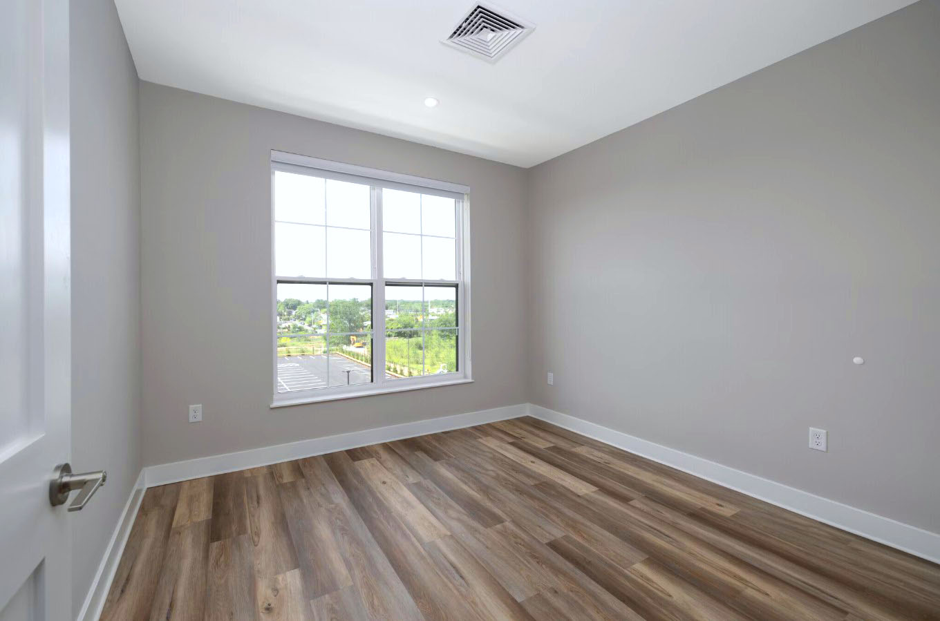 A bright, vacant bedroom at Creekline at Fairfield features wide-plank flooring and a large window providing an expansive view of the surrounding greenery and parking area.