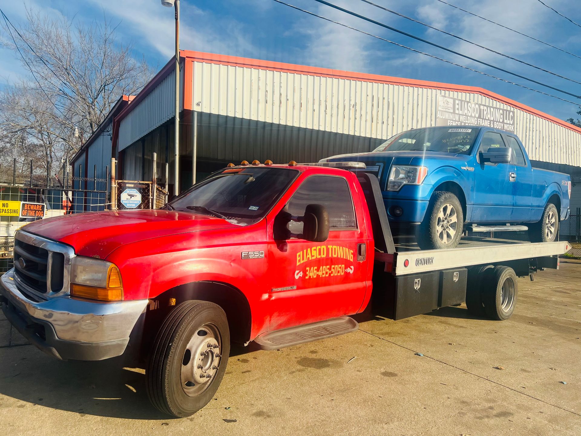 A red Eliasco Towing F-550 flatbed truck transports a blue Ford F-150 pickup truck, parked outside a metal warehouse building labeled Eliasco Trading LLC.