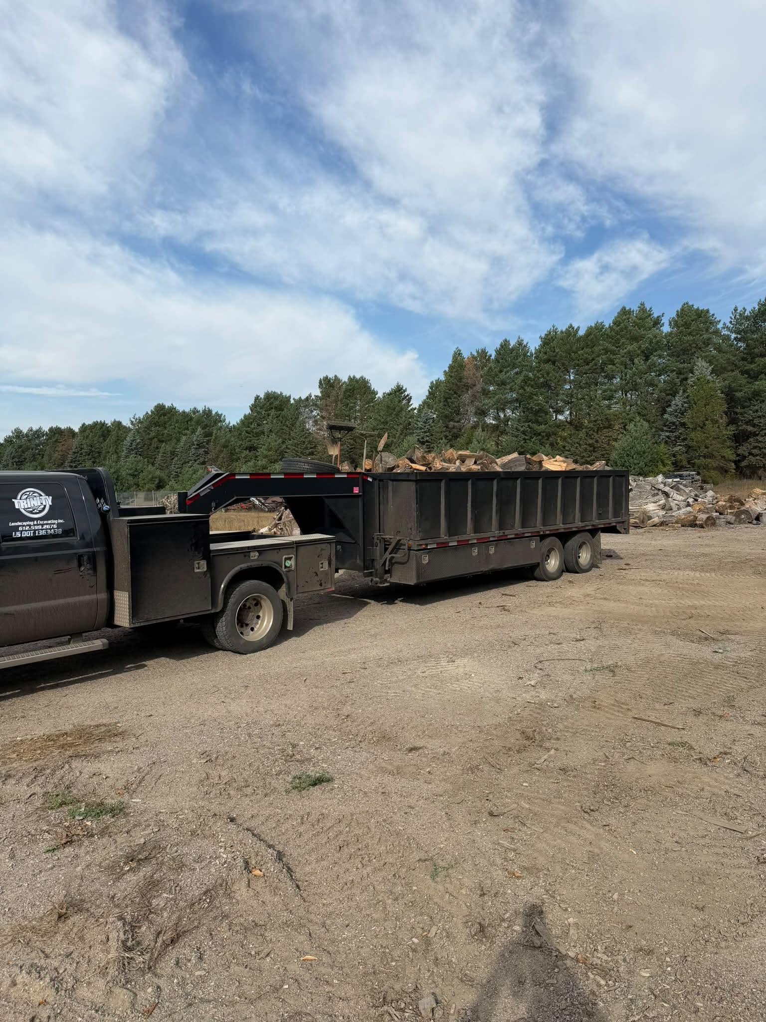 A side view of a heavy-duty black commercial work truck attached to a large, black gooseneck dump trailer filled to capacity with split firewood. The rig is parked in a wide gravel staging area with tall evergreen pine trees under a blue sky with soft white clouds.