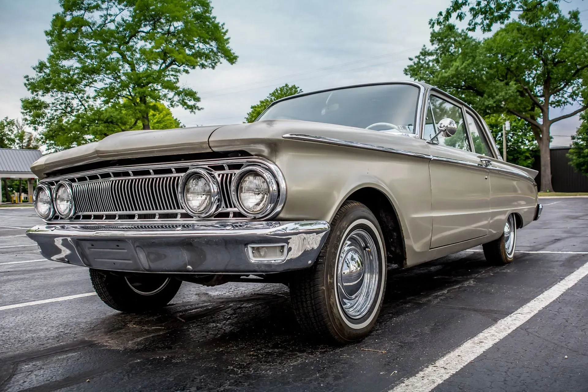 A vintage, light-colored Mercury sedan is parked on a wet asphalt surface in an outdoor parking lot. The car is seen from a low-angle, front-three-quarters view, highlighting its chrome grille, dual headlights, and whitewall tires. Lush green trees and a cloudy sky form the background. The car appears to be in excellent condition, suggesting it is a classic or well-maintained vehicle.