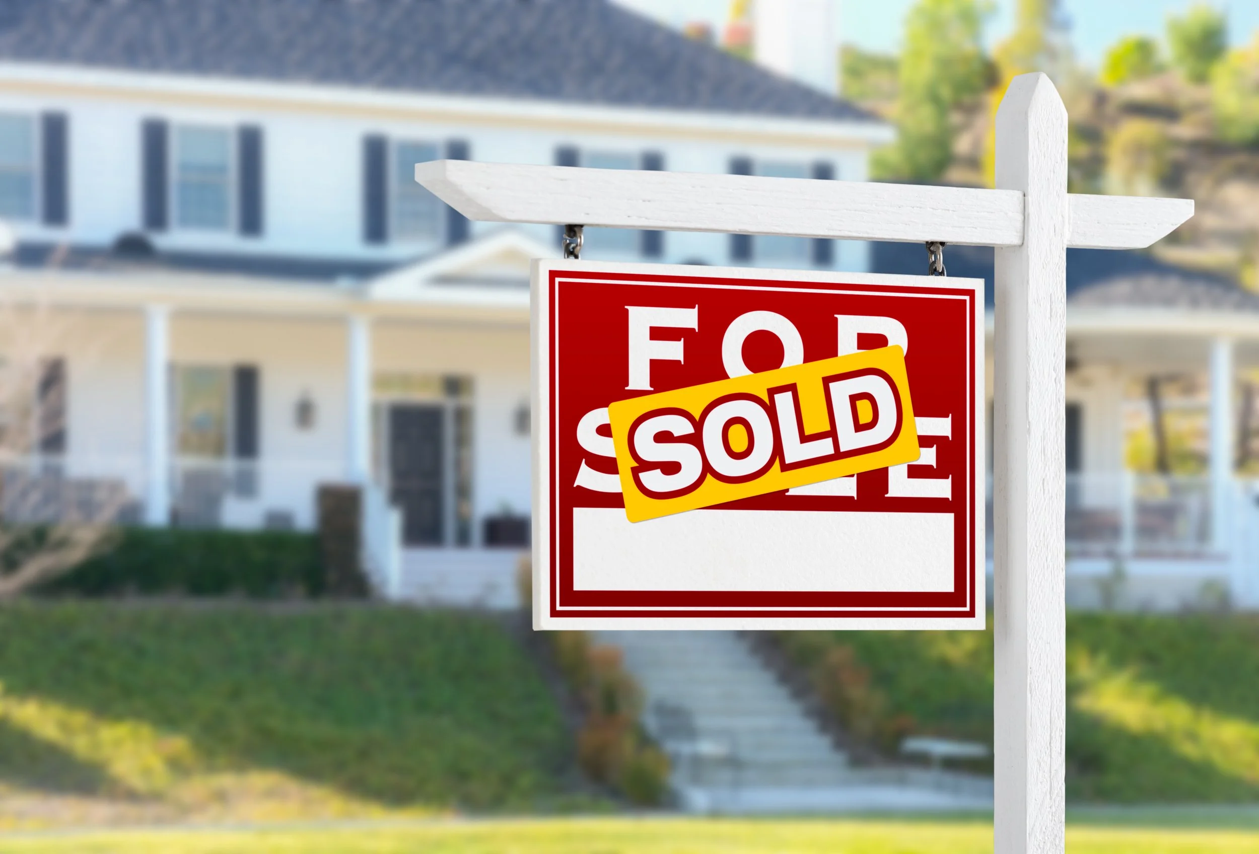 A white wooden post holds a red and white 'For Sale' sign with a yellow 'SOLD' banner across it. In the background, a large white house with dark shutters and a covered porch is visible, surrounded by a green lawn and trees.