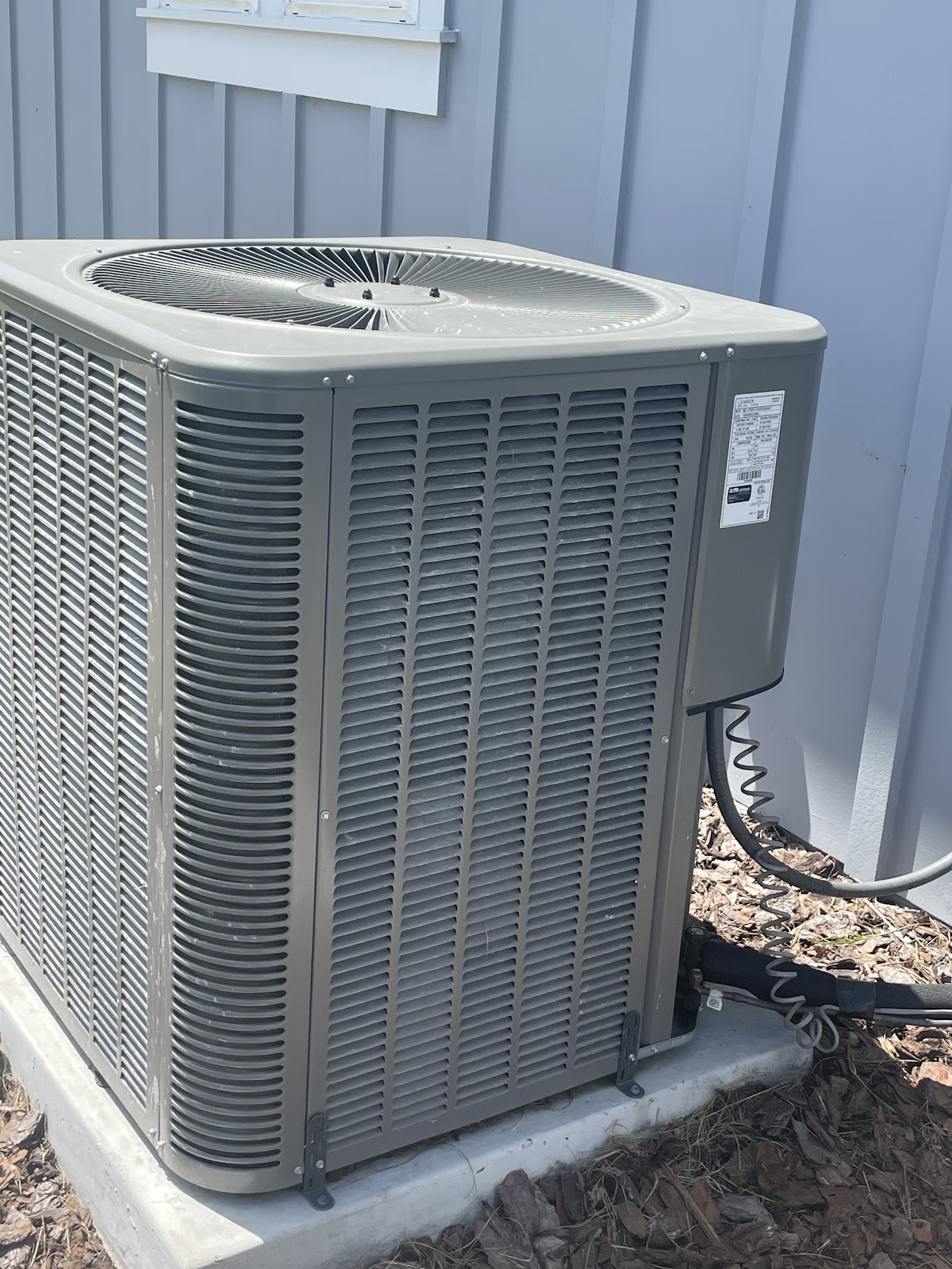 A close-up outdoor shot of a gray air conditioning unit, with a white-trimmed window visible above a 