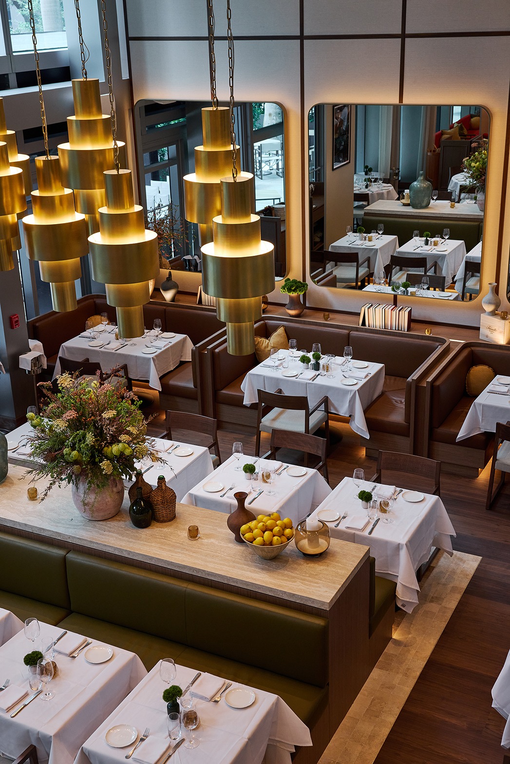 A spacious dining room featuring dramatic gold pendant lights, olive green and brown leather seating, and large mirrors reflecting an elegant, warm atmosphere.