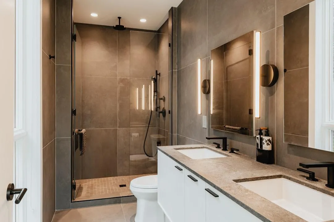 A modern bathroom with a walk-in shower, a double vanity with two sinks, and contemporary lighting fixtures. The walls are tiled in a neutral gray, and the vanity is white with a speckled gray countertop. Black fixtures and hardware complete the sleek look.