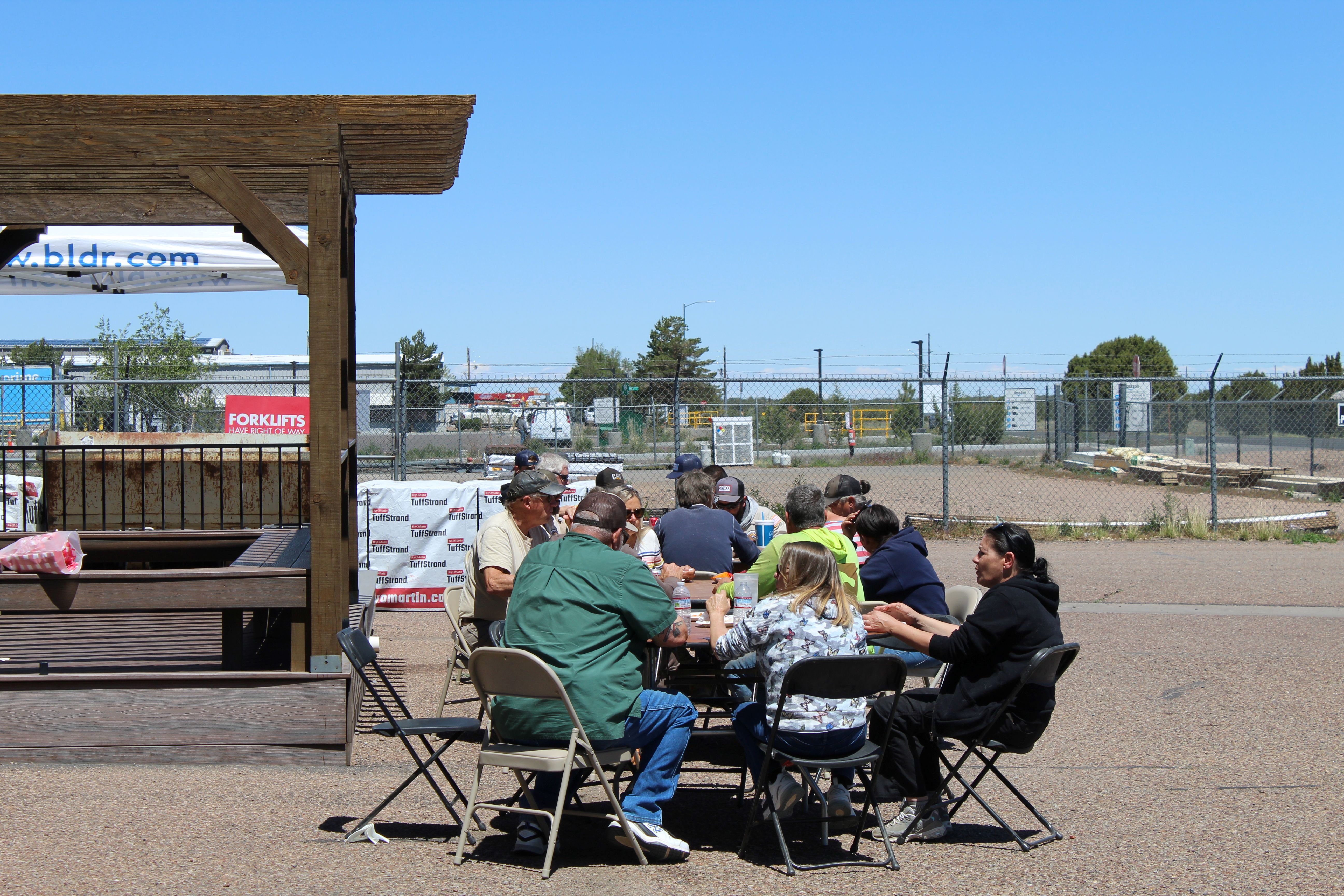 Group of people seated around folding tables outdoors, eating together near a wooden shelter, with a fenced lot, signs, and industrial equipment visible in the background.
