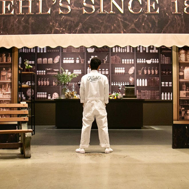 A man dressed in all white stands with his back to the camera wearing a white jacket that says "Kiehl's" on the back