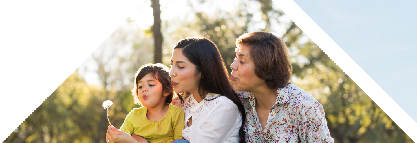 A grandmother, her daughter, and her young granddaughter sitting outside blowing a dandelion together.