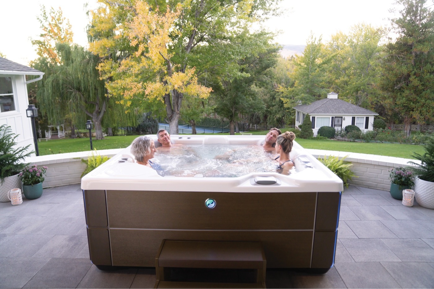 A Hot Spring Highlife Vanguard hot tub (2022) in an Alpine White shell with a Java wood-grain cabinet is shown partially recessed into a raised patio surround on a large residential property. Four adults &mdash; two couples spanning different generations &mdash; are relaxing together in the fully jets-on spa. The unit features the iconic Hot Spring illuminated logo on the front cabinet panel and a matching Java step. The spa is set on a large-format stone tile patio, flanked by potted ferns, flowering plants, and candle lanterns. The lush backyard setting includes mature trees with fall foliage, a well-kept lawn, and a guest house visible in the background.