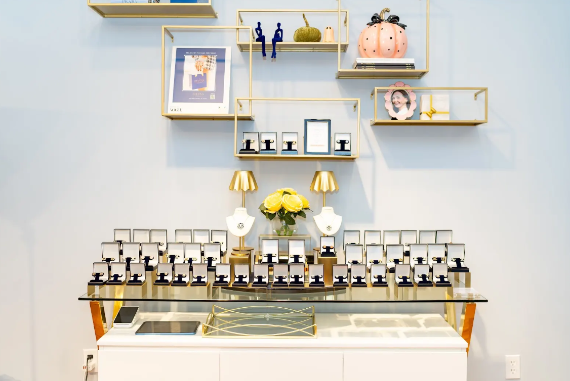 Eliana Curated showcases rows of boxed earrings, pendant necklaces, and gold-tone shelving accented with yellow roses and fall decor. Explore this inviting jewelry boutique in Old Town Alexandria, VA.