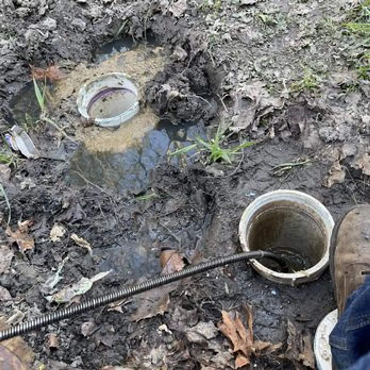 A close-up view of a plumber's tool, a drain snake, being inserted into a pipe opening in muddy terrain. The scene includes a person's boot and leg, indicating a work in progress.