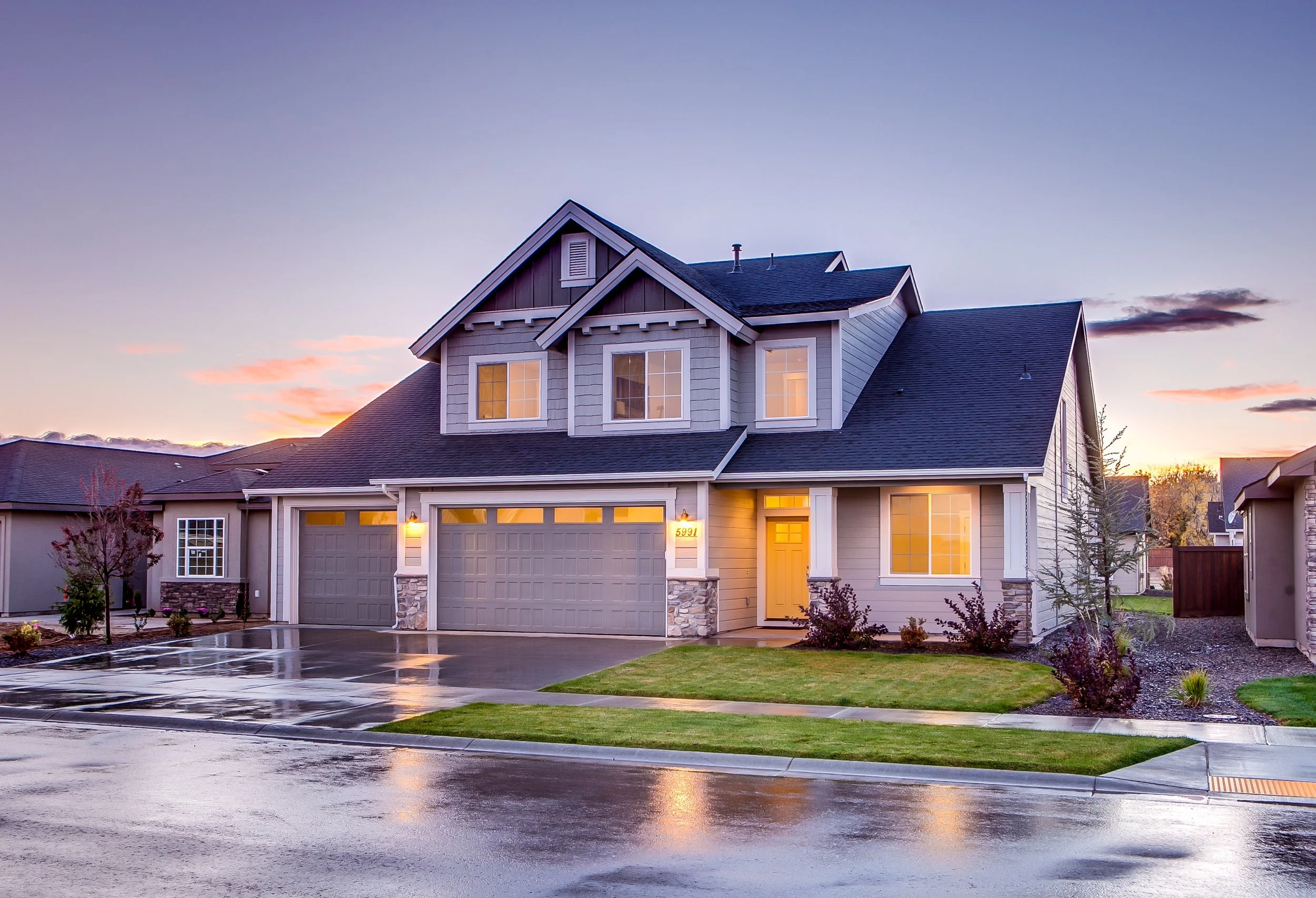 A suburban house with a grey roof and siding is shown at dusk, with warm light emanating from the windows and a lit yellow front door. The driveway and street in front of the house are wet, reflecting the lights and the colorful sky. The sky is a gradient of soft purple and pink, with a few wispy clouds.