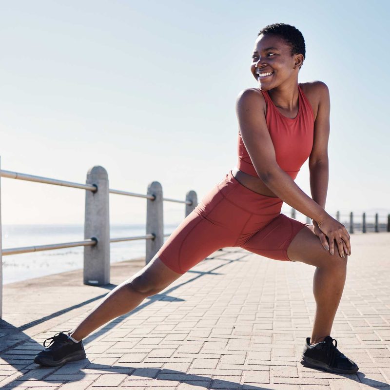 A young Black woman with short hair is stretching her legs while wearing a coral-colored sports bra and matching shorts. She is outdoors by the water, possibly on a boardwalk or promenade, with a railing behind her and the ocean visible in the distance. The sun is shining brightly, casting shadows on the brick-patterned ground.