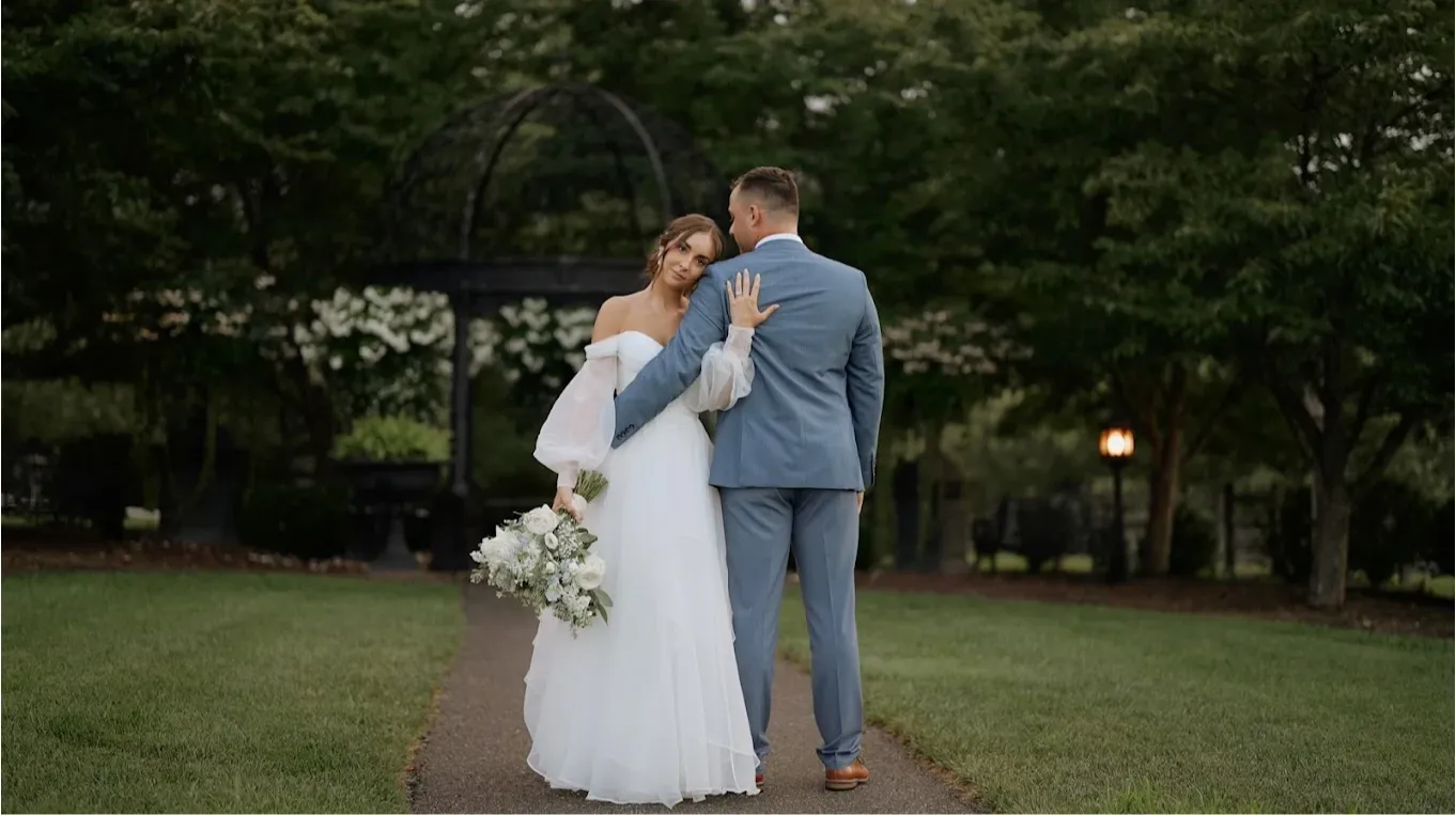 wedding photo of groom standing with his back facing the camera and the bride leaning against him while looking at the camera.