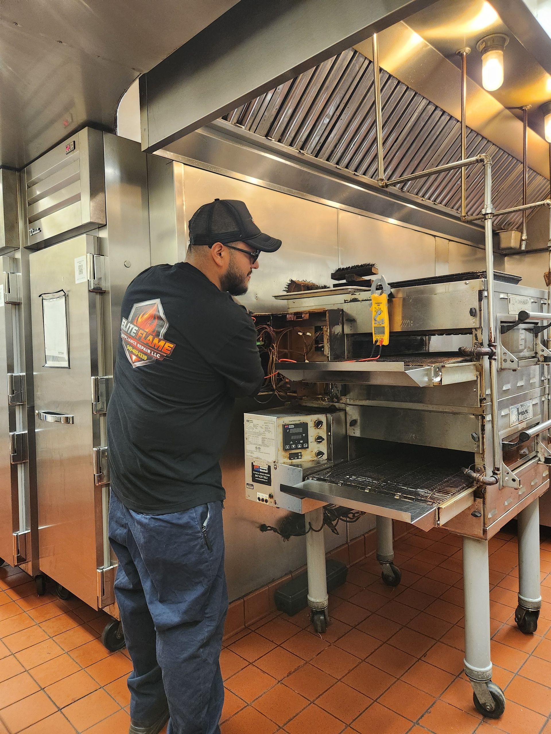 Elite Flame technician inspecting and repairing a commercial conveyor oven inside a restaurant kitch 