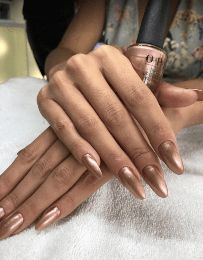 A close-up shot of a woman's hands with perfectly manicured, long, stiletto-shaped nails painted in a metallic rose gold hue. One hand holds a bottle of OPI nail lacquer of the same color. The background is softly blurred, showing a hint of a floral print blouse and a white terry cloth surface, suggesting a salon or spa setting.