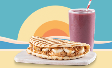 An image of a flatbread filled with fresh bananas, creamy peanut butter on a white kitchen cutting board, behind it is a purple smoothie and a sunset behind that.