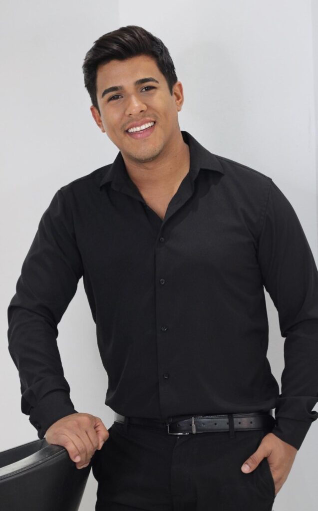 A young man with dark hair and a friendly smile is wearing a black collared shirt and black pants. He is leaning against a black chair with his left arm resting on it. His right hand is in his pocket. The background is a plain white wall.
