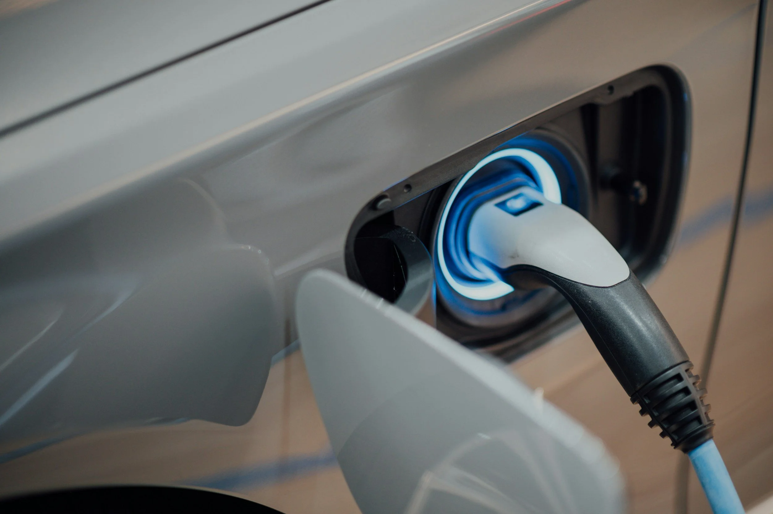A close-up shot shows a silver electric vehicle (EV) being charged. The charging port is open, and a white and black charging cable is inserted, with a bright blue light illuminating the inside of the port.