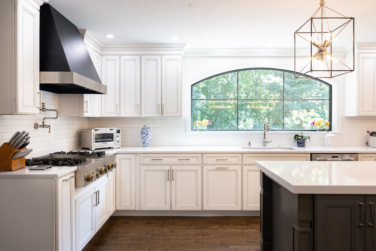 A spacious, modern kitchen with white cabinetry, a large black range hood, and a prominent arched window overlooking a lush green landscape. The countertops are white, and the flooring is dark wood. A kitchen island is partially visible on the right.