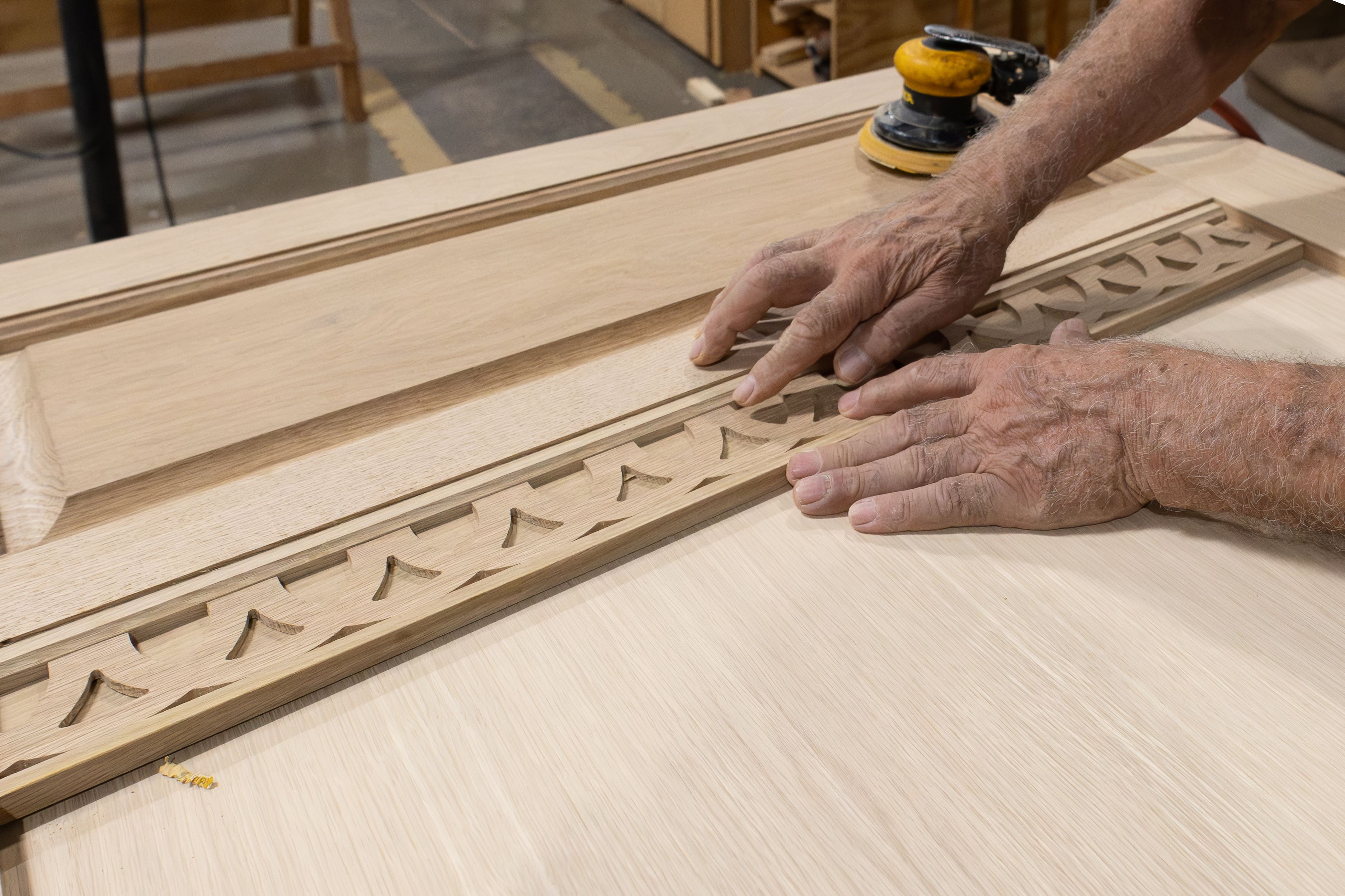 Close-up of hands carefully aligning intricate wooden trim pieces on a door panel during the custom millwork process, with a sander tool visible in the background.