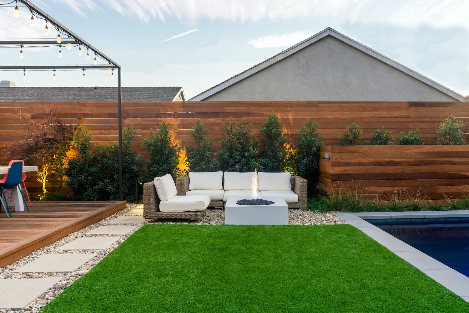 Modern backyard patio with lush artificial lawn, white outdoor sofa, fire table, wood fence, greenery, string lights, and swimming pool.
