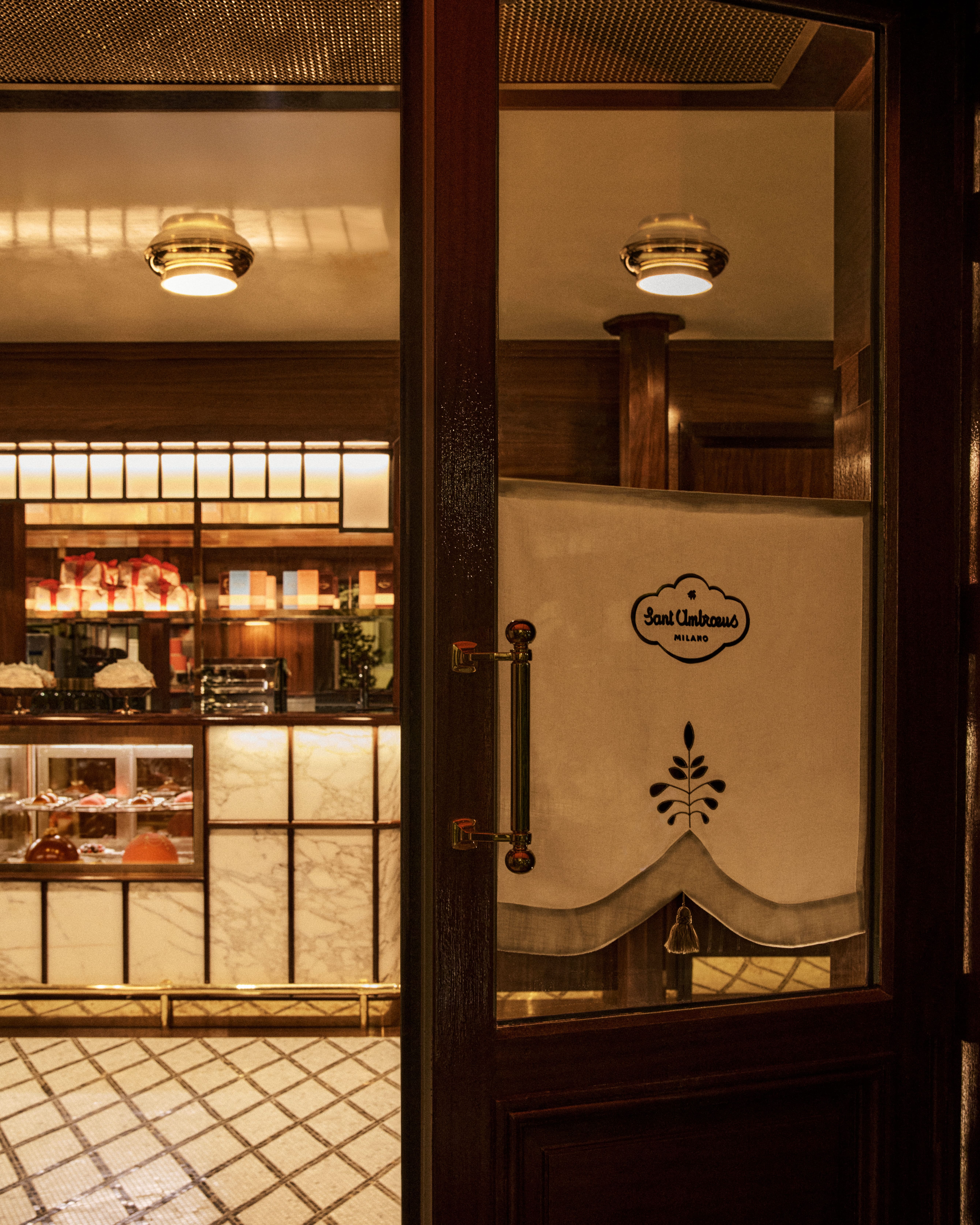 Partially open wooden door with frosted glass bearing the Sant Ambroeus Milano logo, revealing a softly lit pastry counter with marble panels, brass details, and tiled floor inside an elegant café interior.