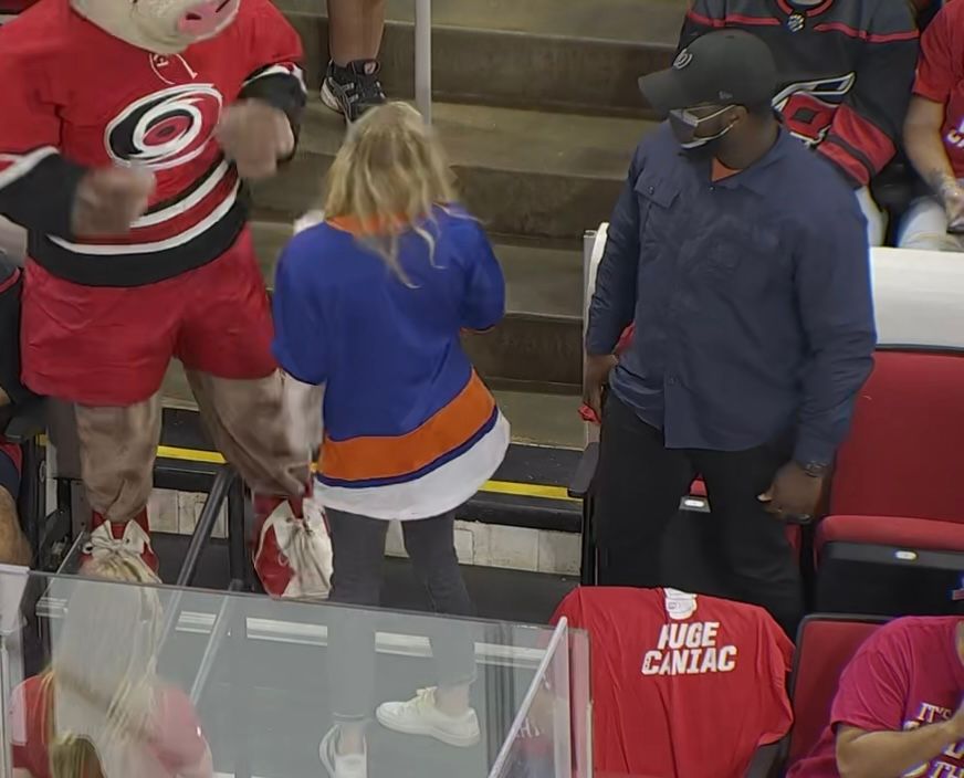 A sports mascot interacts with a woman in a blue jersey near rink-side seats, while a man in dark clothing observes.