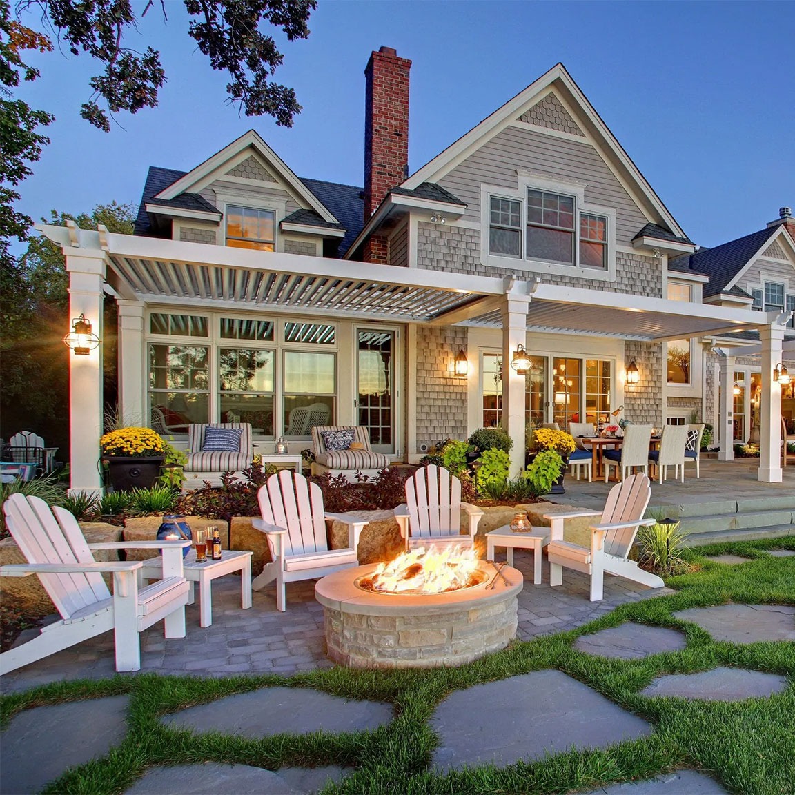 White wood pergola with custom stone gas fire pit, Adirondack seating, and outdoor dining at twilight on a bluestone patio behind a shingle-style home