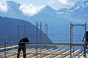 A construction worker assembles a wooden framework atop a scaffold with breathtaking mountain views in the background. The scene captures both the rugged natural beauty of Colorado and the precision of construction work.