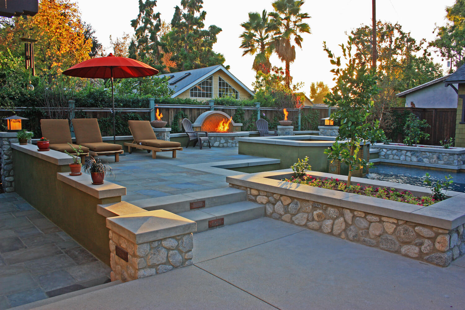 Stone patio garden with lounge chairs, red umbrella, fire pit, raised flower beds, palm trees, and a warm sunset creating a serene retreat.