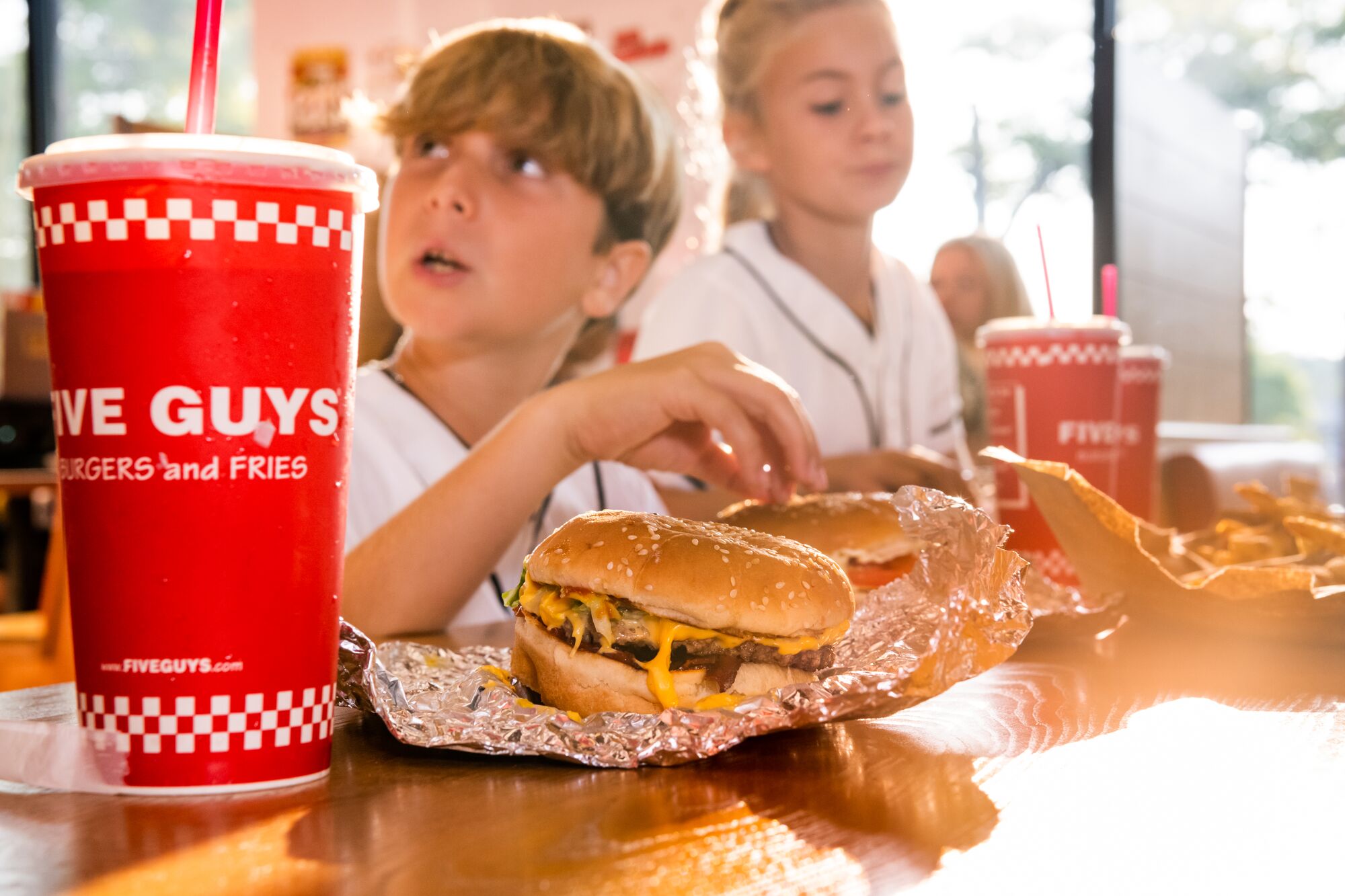 A red Five Guys soft drink cup is the focus in the foreground, as children enjoy their meals in the  Five Guys Thousand Oaks (805)496-0173