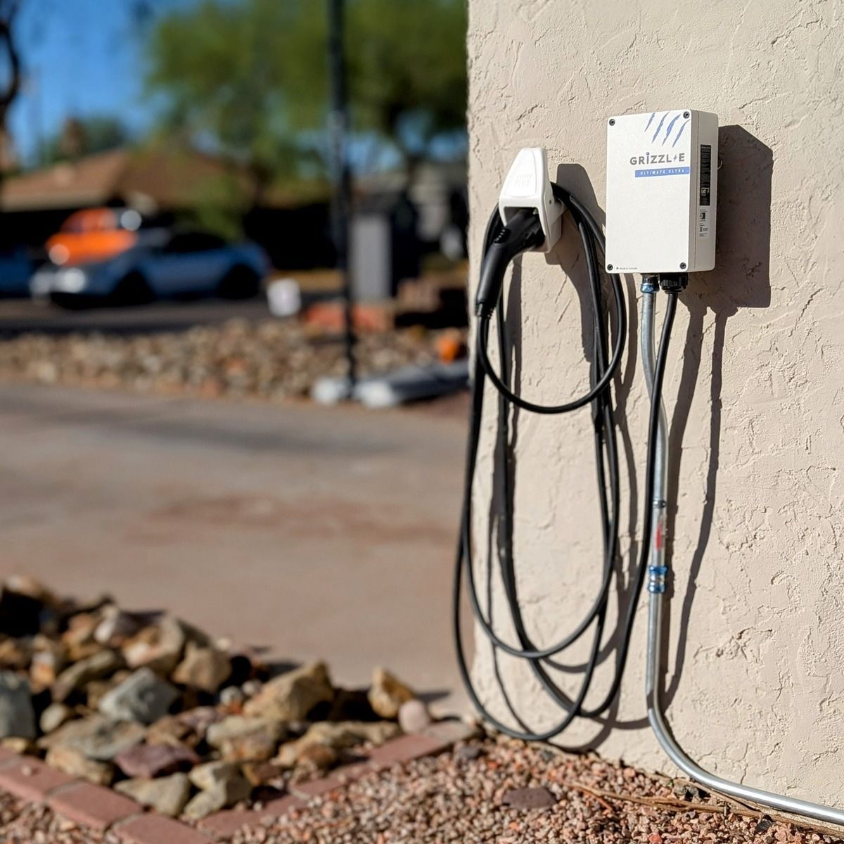 A Grizzl-E electric vehicle charger is mounted on an exterior stucco wall with charging cable hanging neatly beside metal conduit, creating a weather-ready residential charging setup near the driveway outdoors.