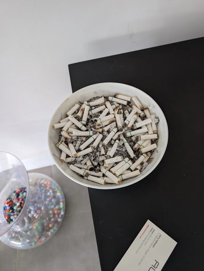 A white ceramic bowl filled with cigarette butts sits on a black surface next to a clear glass container holding colorful marbles. A business card is visible in the lower right corner.