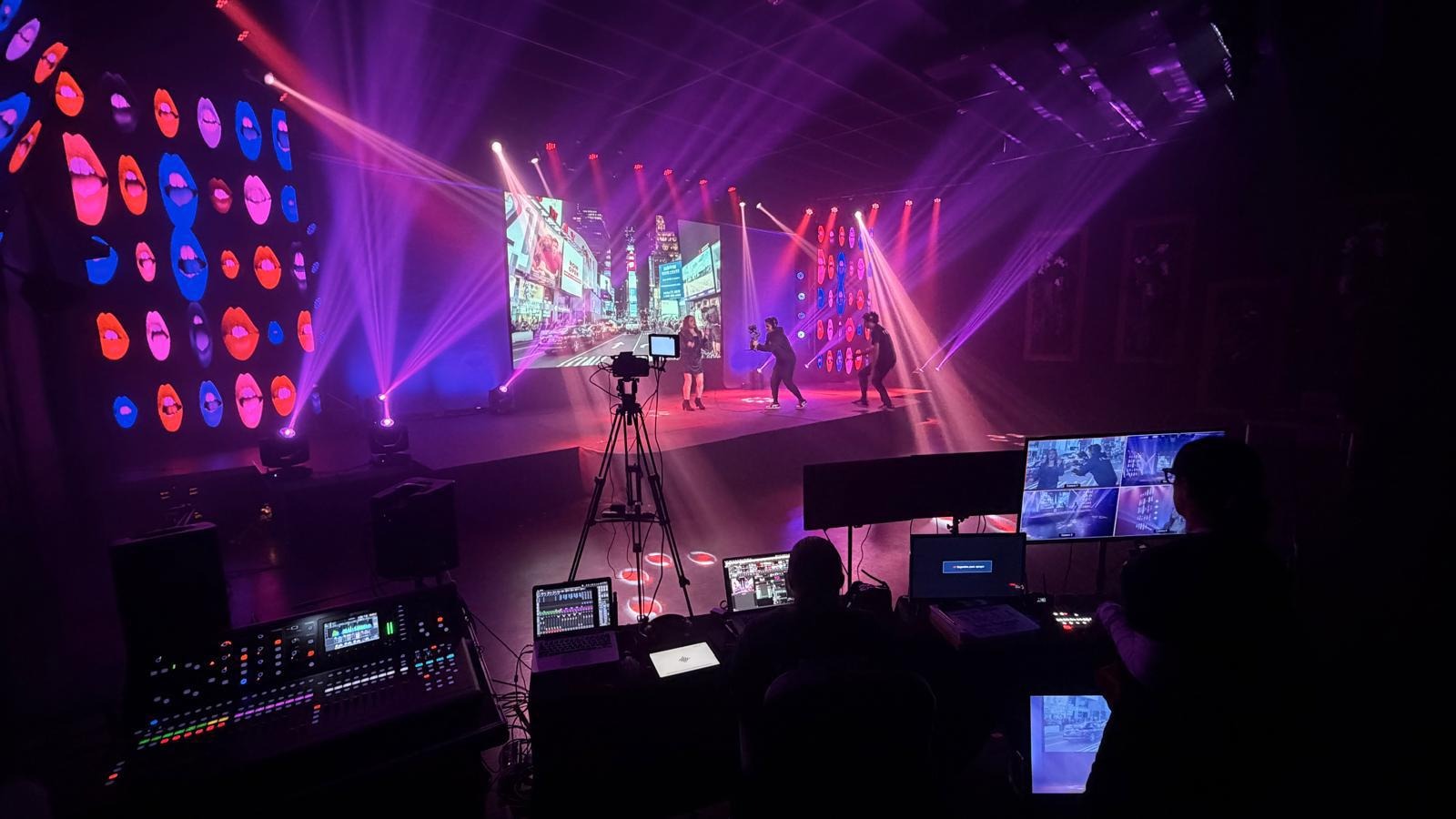 En esta foto te mostramos nuestra cabina de control y producci&oacute;n t&eacute;cnica en plena acci&oacute;n. En primer plano se ven nuestras consolas de audio, monitores y computadoras operadas por nuestro equipo. Al fondo, se aprecia el escenario iluminado con luces rob&oacute;ticas p&uacute;rpuras, donde una cantante y un camar&oacute;grafo realizan una toma frente a una pantalla LED gigante. Como productora de eventos, cuidamos cada detalle en nuestro sal&oacute;n para eventos corporativos. Si buscas el mejor alquiler local para eventos, esta es la calidad t&eacute;cnica que te ofrecemos.