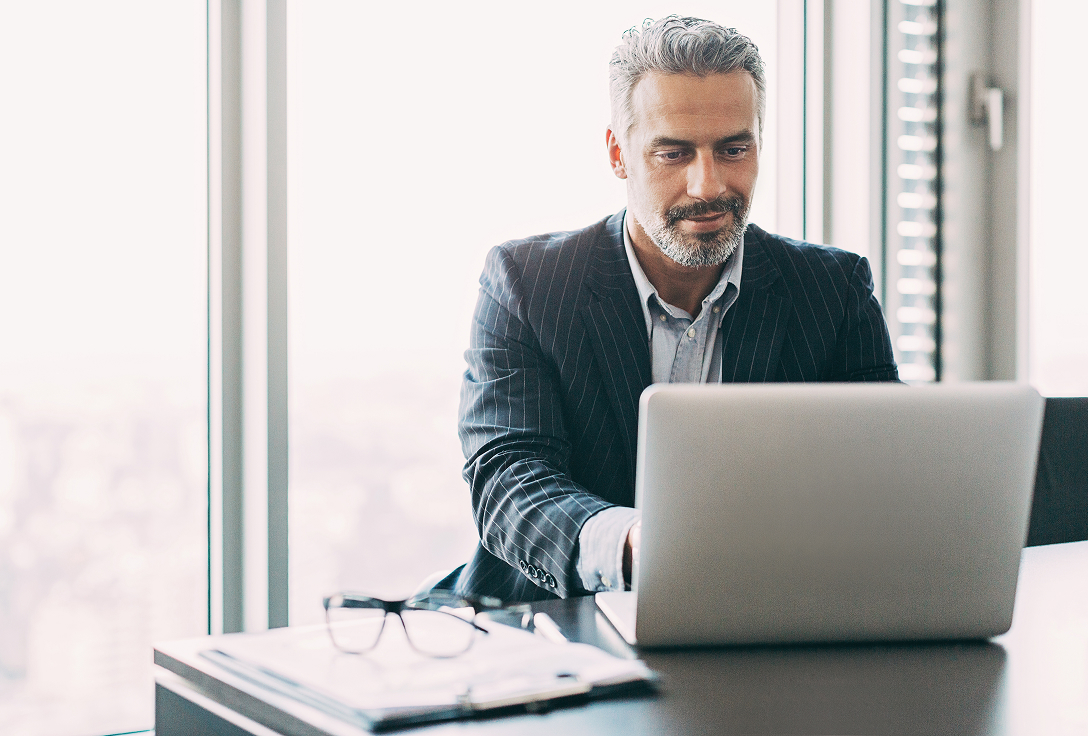 Businessman in a suit working on a laptop at an office desk with eyeglasses and documents. He looks focused, with city skyline visible outside.