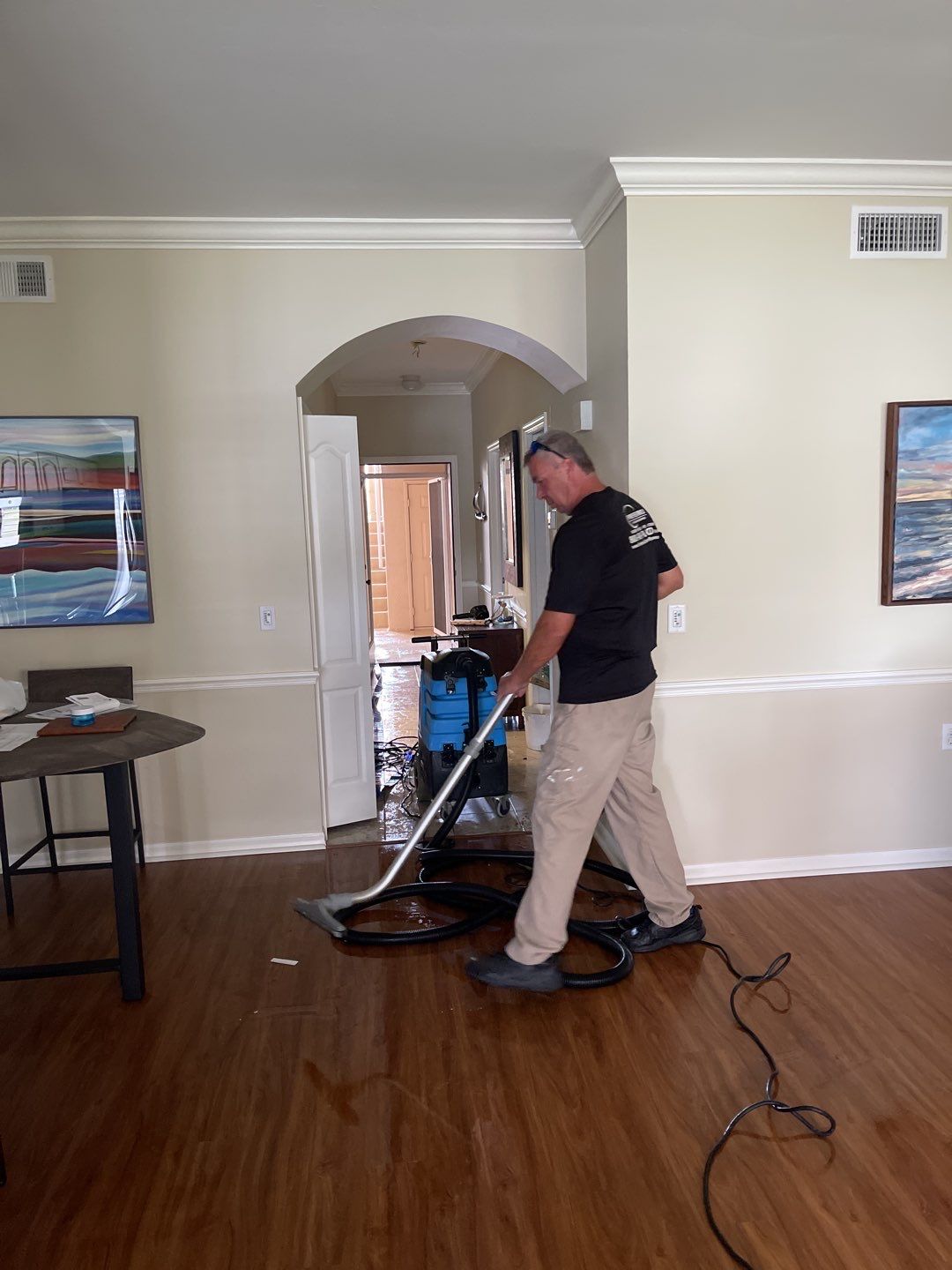 A man wearing a black t-shirt and tan pants is using a wet vacuum cleaner to remove water from a flooded room. The wet vacuum machine is blue and black. He is standing on a wooden floor which is covered in water. Through an arched doorway, another room is visible with more water on the floor. Artwork hangs on the walls of the room, and a round table with chairs is to the left.
