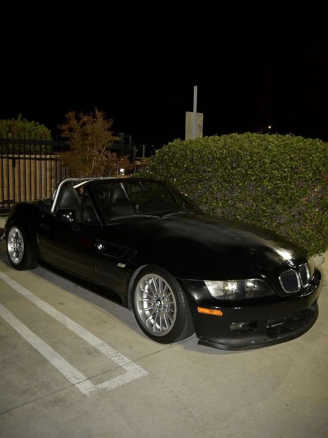 Black BMW roadster parked at a nighttime car meet beside trimmed hedges and illuminated parking spac 