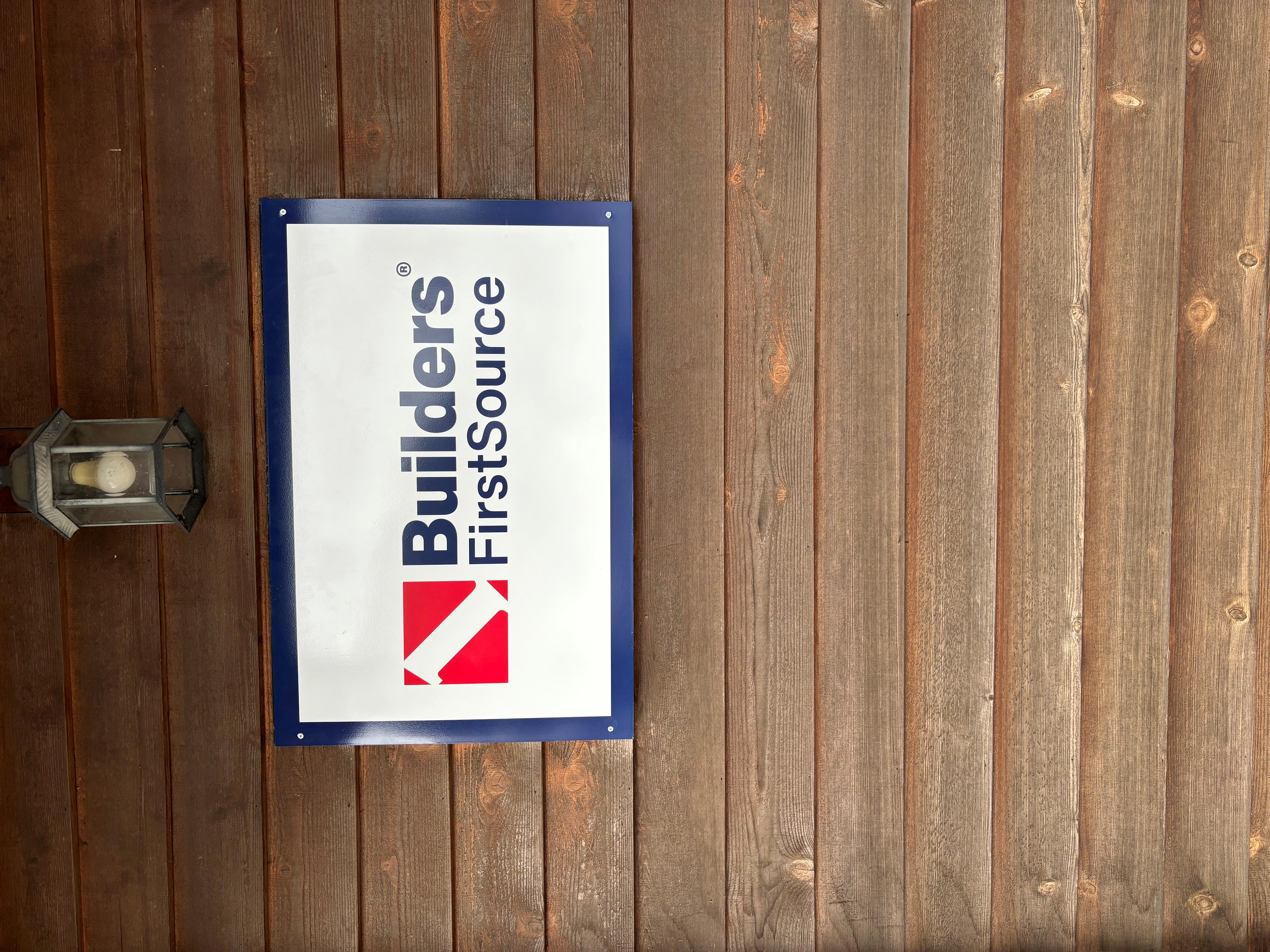 A Builders FirstSource logo sign mounted on a wood-paneled wall, accompanied by a classic outdoor lantern light fixture, creating a rustic and professional aesthetic.