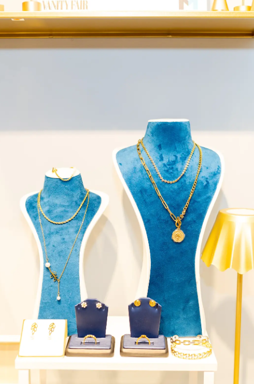 Layered gold-tone necklaces with coin and pearl accents stand out on blue velvet displays at Eliana Curated. Earrings, rings, and bracelets complete a jewelry display in Old Town Alexandria, Virginia.