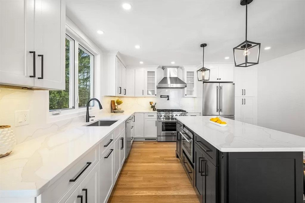 A spacious, modern kitchen with white shaker cabinets, a large island with a black base, a stainless steel refrigerator, and a professional-grade gas range. The countertops are white quartz with gray veining, and the flooring is light wood. Black hardware and pendant lights add a contrasting touch.