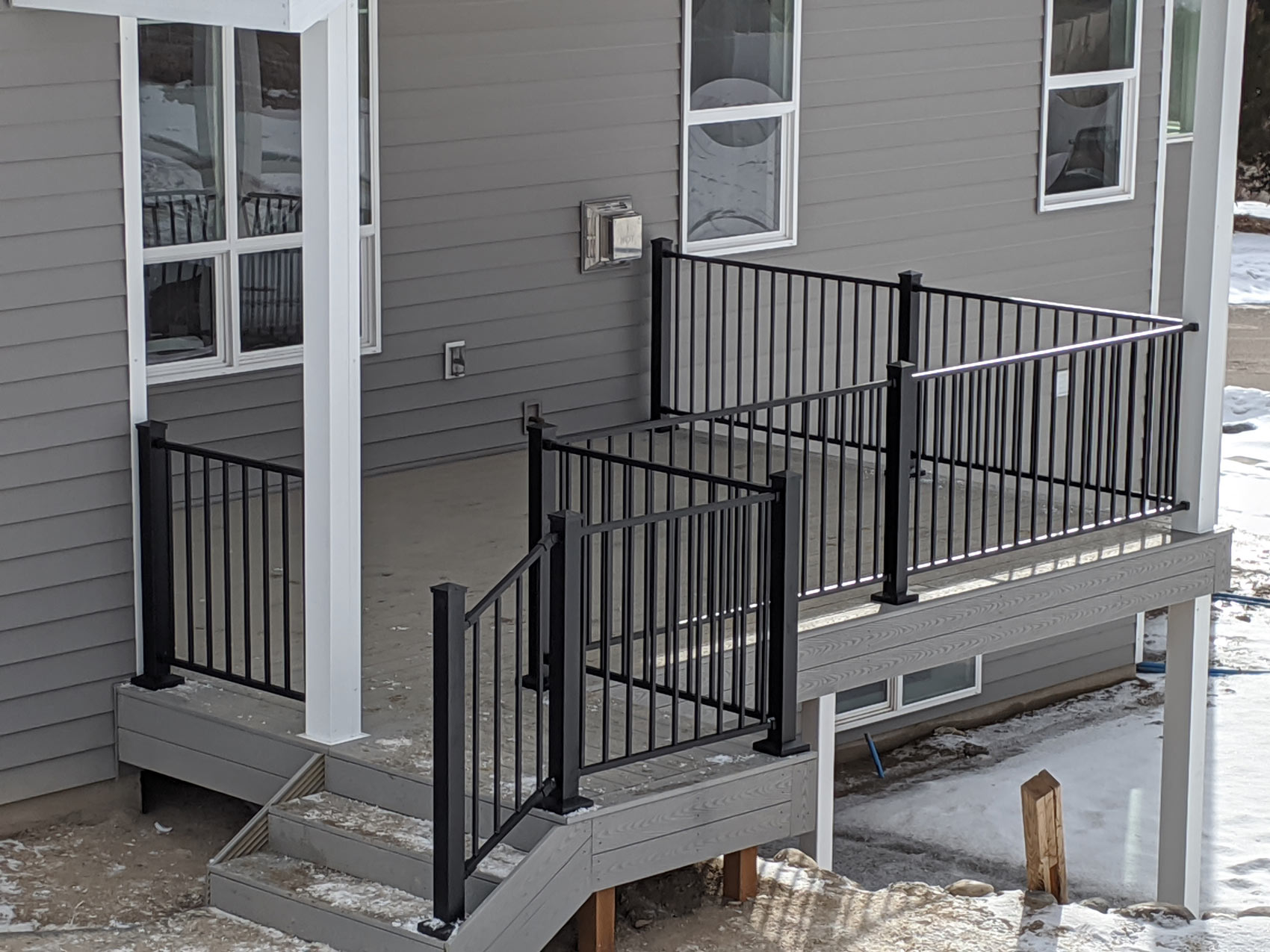 Expand your outdoor living possibilities with this custom grey composite deck by DeckPro LLC. This design provides a safe and stylish transition from your home to your backyard, even in snowy conditions, thanks to durable, low-maintenance composite materials. The contrasting black metal railings and wide steps offer a modern touch while ensuring safe and easy access from the house to the natural terrain below.