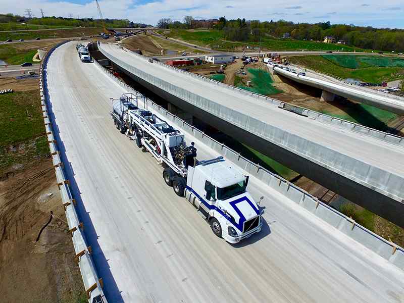 Elevated aerial shot looking along a newly constructed concrete highway bridge deck. A large white Volvo truck towing a heavy DSI specialized grinding rig is parked in a closed lane, with an adjacent unopened bridge span running parallel on the right.