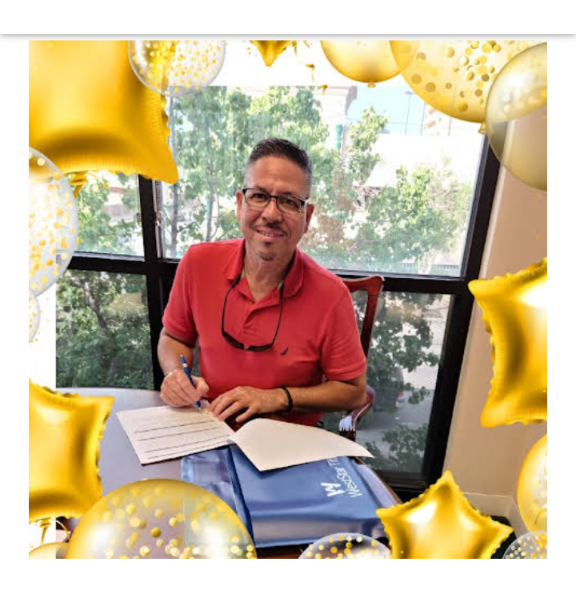 A man wearing glasses and a red polo shirt is sitting at a desk and signing a document. He is surrounded by gold balloons and confetti.