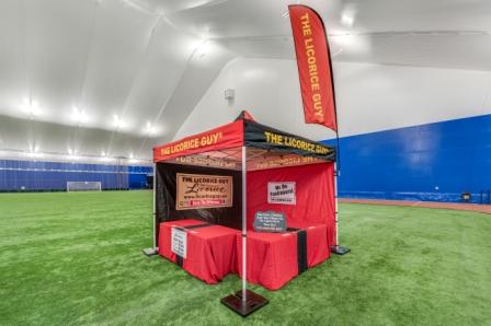 The Licorice Guy's portable event setup, featuring the branded red and black pop-up tent with feather flags and product displays, ready for an event on an indoor turf field.