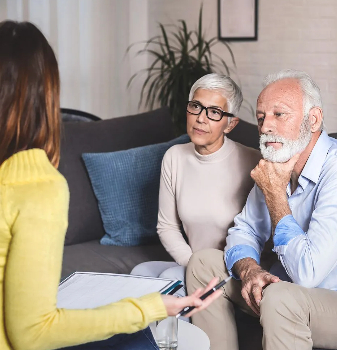 An organizer or move manager sitting with a clipboard during a consultation with an elderly couple on a gray couch in a living room.
