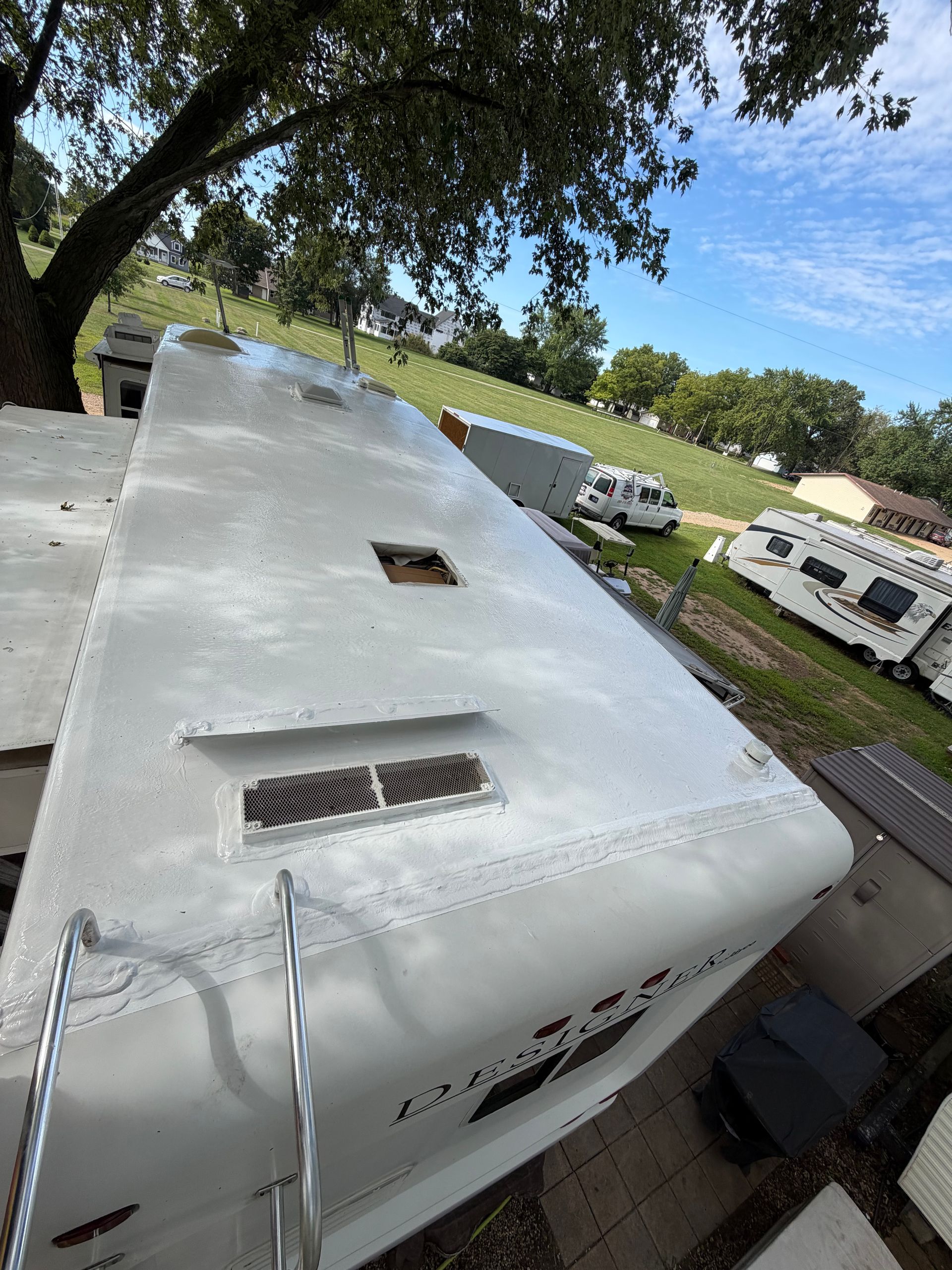 RV roof coated with a fresh white protective surface, showing vents, openings, seams, ladder rails, and repaired areas, with other recreational vehicles, service vans, lawns, and trees visible nearby outside.
