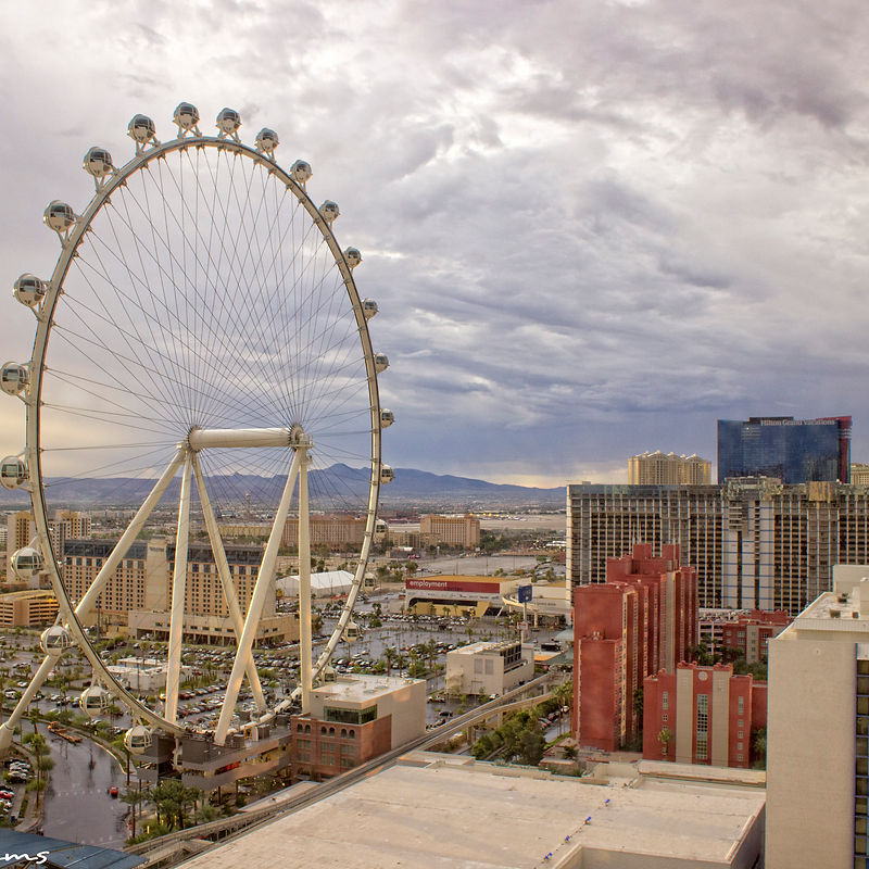 A wide shot of the Las Vegas Strip with the High Roller observation wheel prominently featured in the foreground under a dramatic cloudy sky. Numerous hotels and casinos, including the Hilton Grand Vacations, are visible in the background, along with a large parking lot filled with cars and a monorail track.