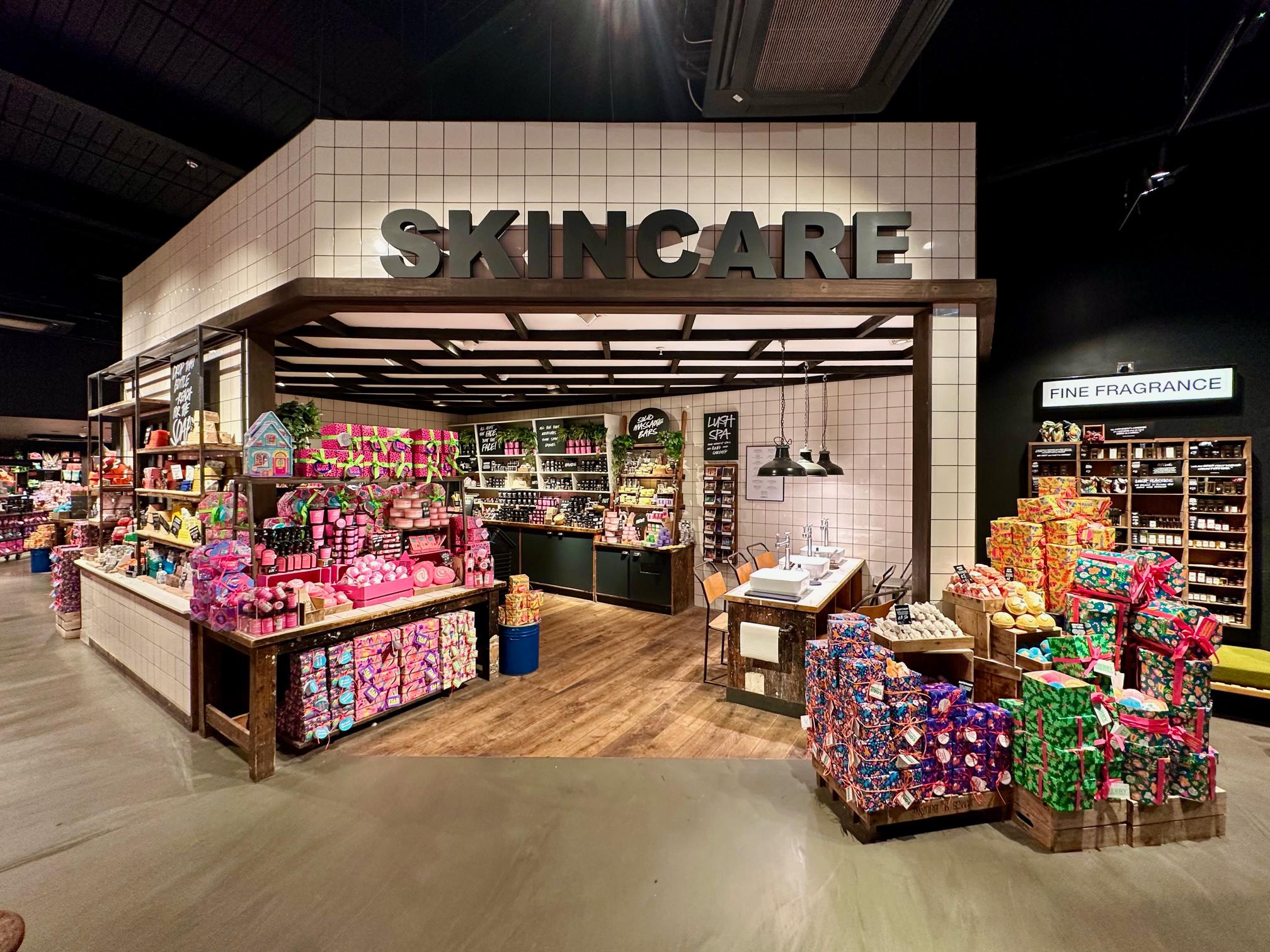 A smaller building with a large black sign saying skincare. On both sides of the entrance are large piles of brightly coloured gifts. Inside the building are three white sinks and a wall full of pots and other products.