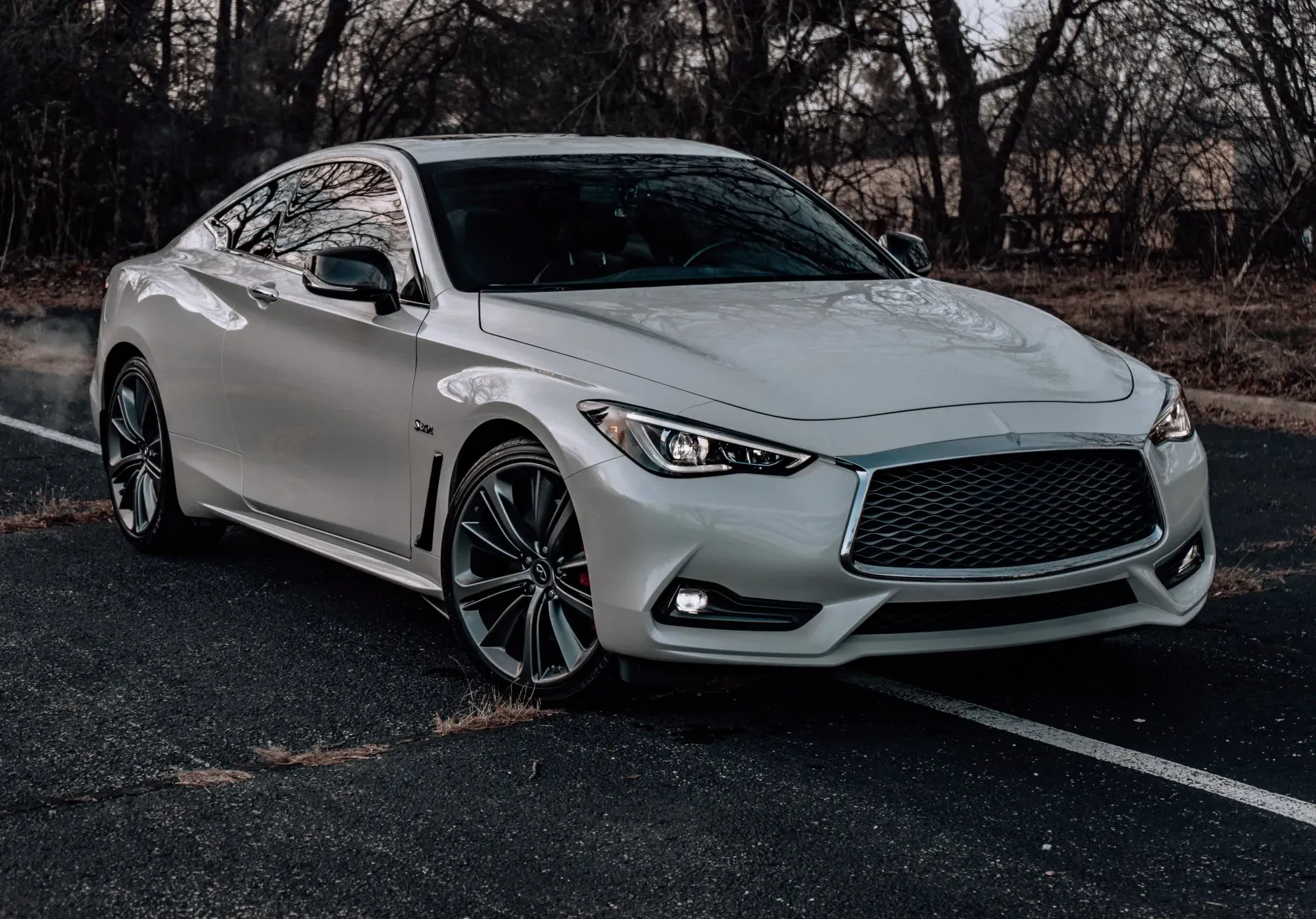 A white Infiniti Q60 coupe is parked on an asphalt road with a backdrop of bare trees and a cloudy sky. The car has a sleek, sporty design with a prominent grille and sharp headlights. The wheels are dark gray with a multi-spoke design, and red brake calipers are visible. The car is angled towards the viewer, with a slight motion blur on the left side, suggesting it might be starting to move or has just stopped.