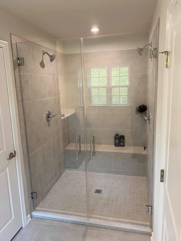 A modern, spacious walk-in shower with a glass enclosure, featuring two shower heads, a built-in bench, and tiled walls and floor. Two bottles of shampoo are visible on the bench.